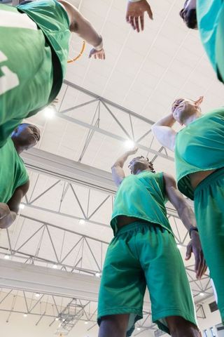 Diverse basketball team huddling under hoop in gymnasium wearing green uniforms