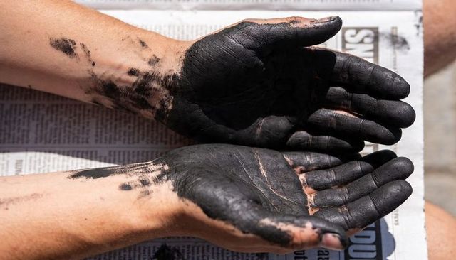 Sunlit hands covered in black charcoal pigment holding palms up on newspaper closeup