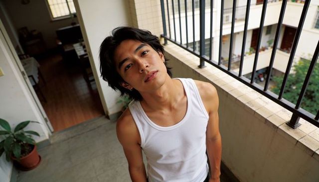 Asian man leaning on balcony railing wearing white tank top, urban courtyard portrait