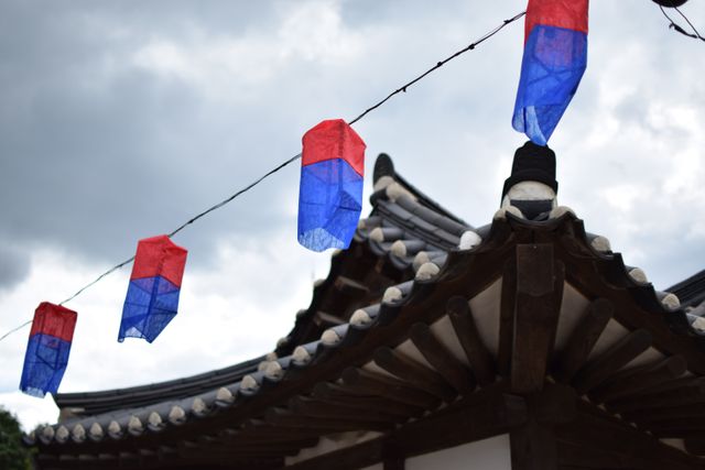 Traditional blue and red lanterns are hanging on string over an ancient, wooden Asian roof with intricate design. Useful for illustrating cultural festivals, traditional architecture, or heritage tourism. Suitable for use in travel brochures, educational materials, and blog posts about Asian culture and traditions.