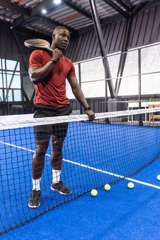 African american padel player holding racket leaning on net on indoor blue court