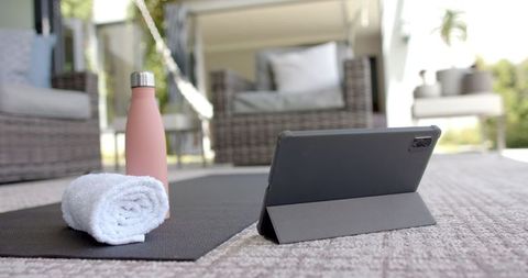tablet resting on yoga mat with nearby towel and water bottle indicates a tranquil workout space, ideal for yoga enthusiasts practicing at home. pleasant outdoor setting with comfortable furniture enhances inviting atmosphere, making it ideal for fitness brands, home wellness advertising, or technology in mindfulness practices.