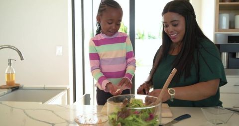 African american mother and daughter preparing fresh salad together in bright kitchen
