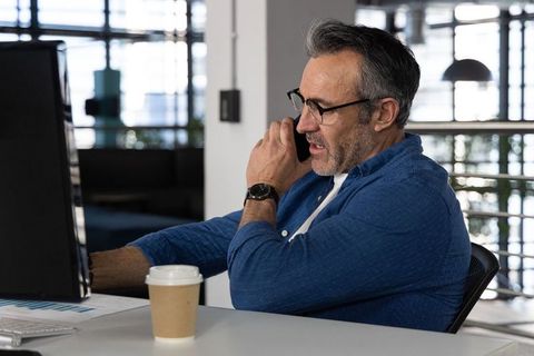 Middle-aged professional talking on smartphone at desk with monitor, coffee and charts in modern off