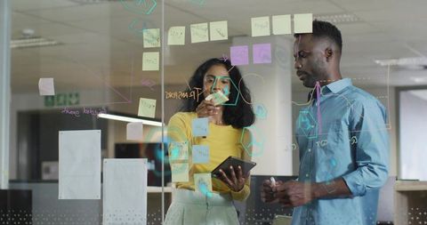 Planning team collaborating on glass wall with sticky notes and tablet in modern office