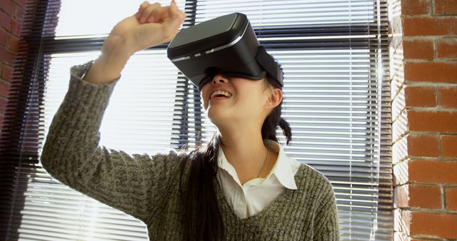 Young woman wearing VR headset, showing excitement while using virtual reality technology indoors. Sunlight streaming through window blinds, with brick wall in background. Ideal for illustrating concepts related to modern technology, virtual adventures, innovation, and immersive experiences.