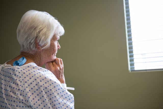 Elderly woman in hospital gown standing near window, appearing deep in thought. Ideal for use in healthcare, medical, and senior care contexts, illustrating themes of aging, patient care, and medical treatment.
