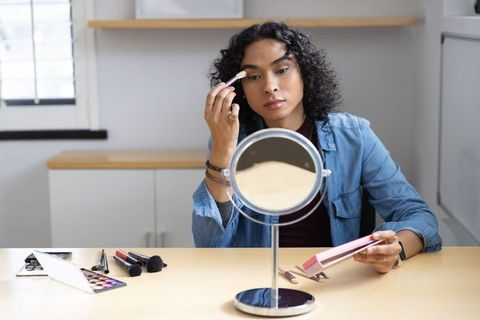 Non-binary person applying eyeshadow at desk with mirror and makeup palette