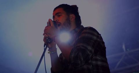 Male vocalist singing into vintage microphone wearing plaid shirt and bracelets, stage lens flare