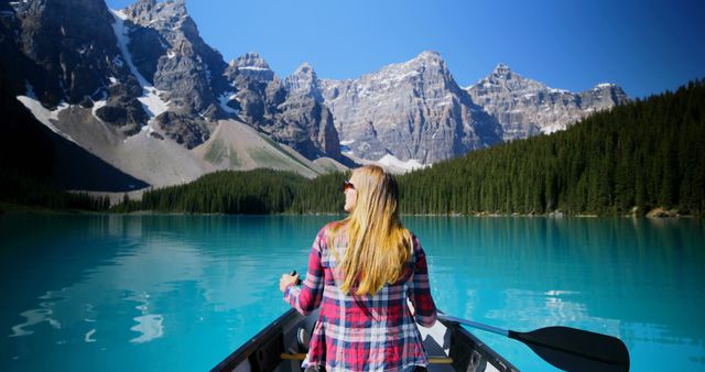 A woman in a plaid shirt is paddling in a canoe on a pristine mountain lake, surrounded by majestic peaks under a clear blue sky. This image can be used for themes related to outdoor adventures, travel destinations, nature retreats, and relaxation. Perfect for websites or magazines focusing on tourism, nature experiences, and motivating people to explore the outdoors.