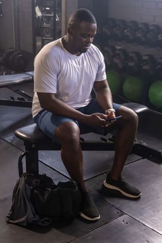 African man sitting on weight bench using smartphone after workout in moody gym