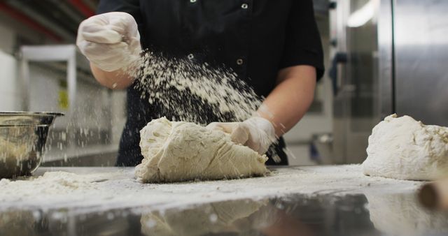 Hands of a baker wearing gloves, kneading dough on a floured surface in a professional kitchen setting. Flour is being sprinkled over the dough and table. This image is perfect for use in culinary, bakery, food industry, cooking courses, and professional kitchen themes.
