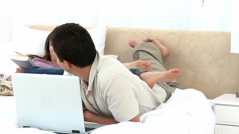 Couple Relaxing on Bed Smiling with Laptop and Book