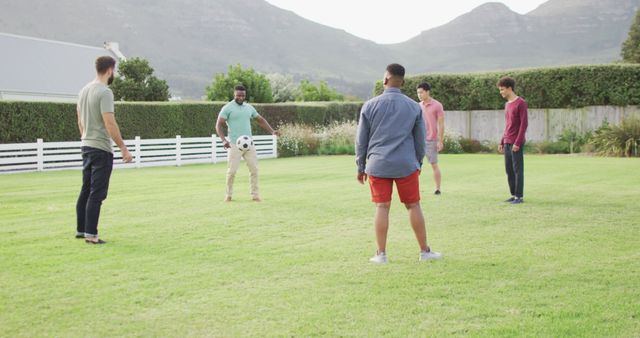 Group of five adult male friends casually playing soccer on a well-groomed grassy field surrounded by nature. Men are standing in a circle, engaged in a friendly game, under clear and sunny skies with mountains in the background. Ideal for use in advertisements promoting outdoor activities, sports equipment, men's fashion, or lifestyle brands focusing on friendship and social bonding.
