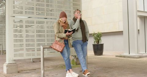 Mature couple leaning on rail checking notebook and sipping coffee in urban plaza