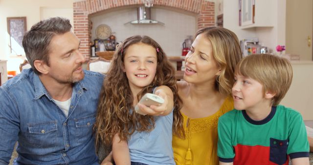 Picture of a happy family, including parents and two children, enjoying TV time together in a cozy living room. The daughter is holding the remote control while the family engages warmly. Perfect for advertisements and articles about family bonding, leisure activities, and modern family life.