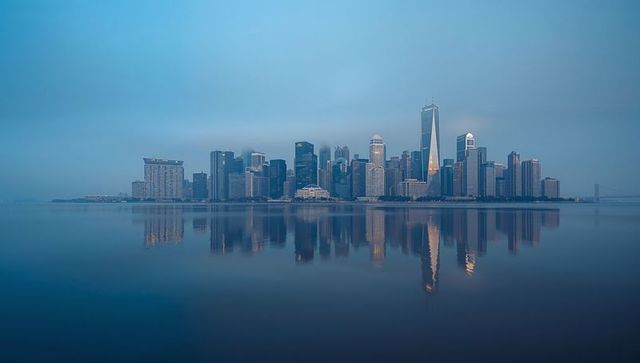 Dawn skyline reflecting on calm harbor waters with slender glass tower and city lights