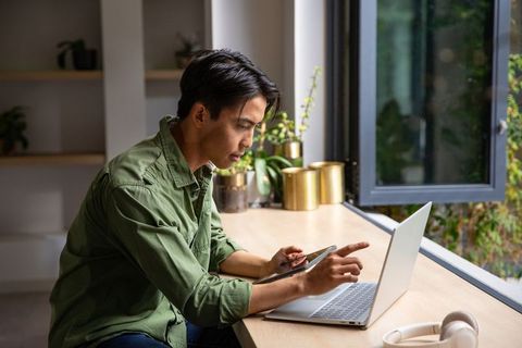 Young asian man working from home on laptop and tablet at sunlit window desk