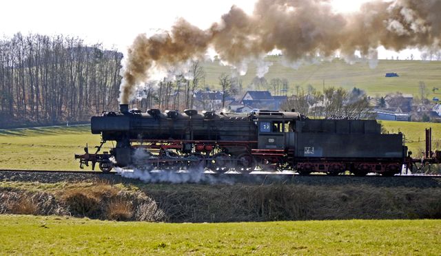 Historic steam train puffing through a rural countryside, emitting thick smoke. Ideal for promoting travel, showcasing historical transport, illustrating rural landscapes, and evoking a feeling of nostalgia. Useful for websites and marketing materials related to railway travel, history, and classic engineering.