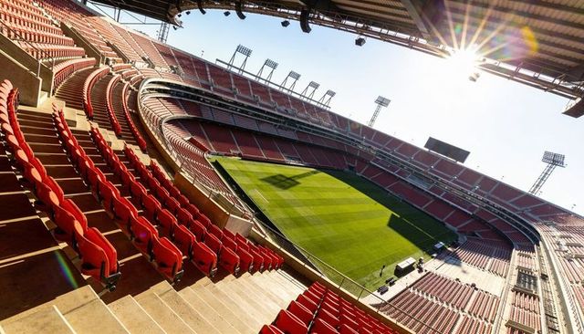 Curving red seats sweeping over sunlit green football pitch in modern stadium bowl