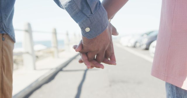 Hands of diverse senior couple walking on sunny promenade by the sea holding hands. Retirement, romance, summer, vacations and senior lifestyle, unaltered.
