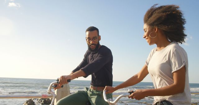Romantic diverse couple riding bikes and smiling on sunny beach, copy space. Summer, vacation, transport, hobby, romance, love, relationship, free time and lifestyle, unaltered.
