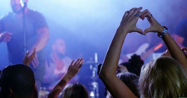 A diverse crowd enjoys a live music performance, with a person making a heart shape with their hands, with copy space. Fans show their appreciation and love for the band playing on stage, creating a vibrant concert atmosphere.