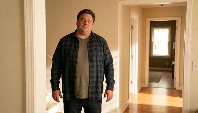 Man standing in sunlit residential hallway wearing plaid shirt and olive t-shirt, pensive