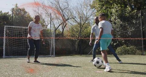 Family playing soccer on backyard turf, child dribbling toward goal with parents