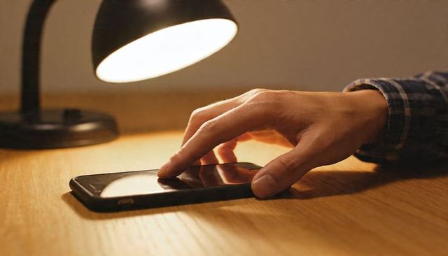 Right hand tapping smartphone on wooden desk under warm lamp light, closeup reflection