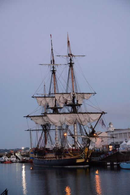 Historic sailing ship docked at a harbor during twilight. Featuring tall masts and billowing sails, this maritime scene captures the beauty of nautical heritage. Ideal for use in historical articles, maritime enthusiasts, travel brochures, and educational materials about seafaring history.