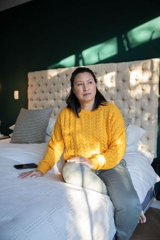 Mature woman sitting on bed edge in mustard sweater, contemplative in warm morning light
