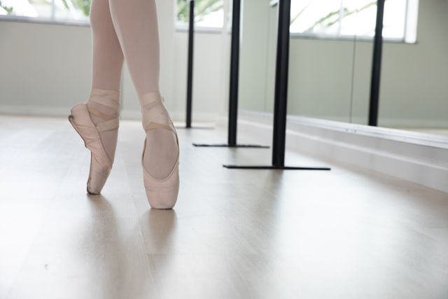 Legs of Caucasian female attractive ballet dancer warming up and practicing in a bright ballet studio, standing on her toes in ballet shoes. Focused on her exercise, preparing for a class.