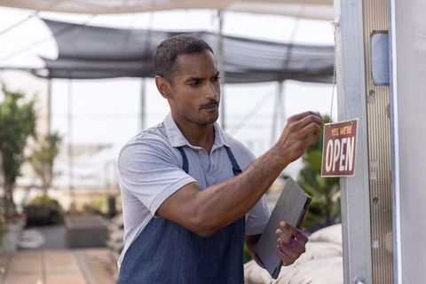 Indian business owner hanging open sign while holding tablet in greenhouse plant nursery