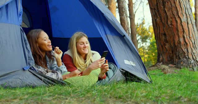 Two women are reclining inside a blue tent in a forest, looking at a smartphone together. The setting suggests they are enjoying a camping trip surrounded by trees and green grass. This image can be used for themes related to outdoor adventures, digital detox, technology, nature lovers, weekend getaways, or bonding time among friends.