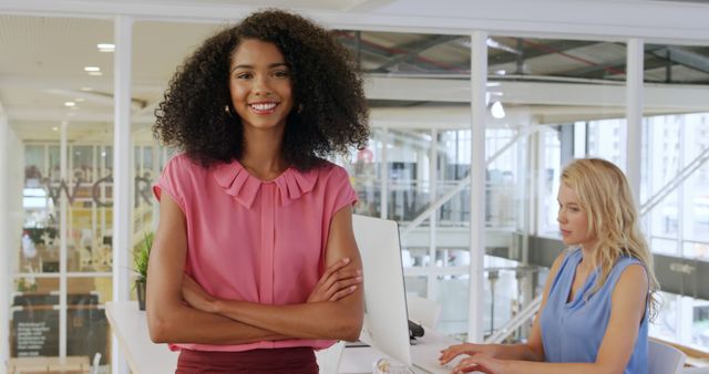 Businesswoman standing confidently with a smile in a modern office environment, while her colleague works on a laptop in the background. This image can be used for business-themed websites, corporate blogs, presentations, and advertisements emphasizing teamwork, professionalism, and modern workspaces.