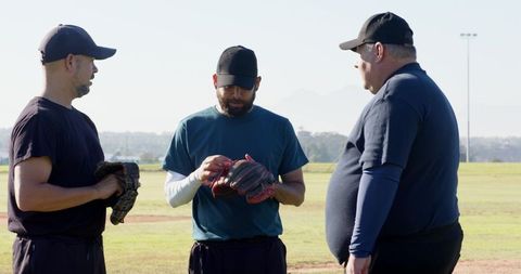 Baseball teammates inspecting glove during practice on sunlit field with coach discussion