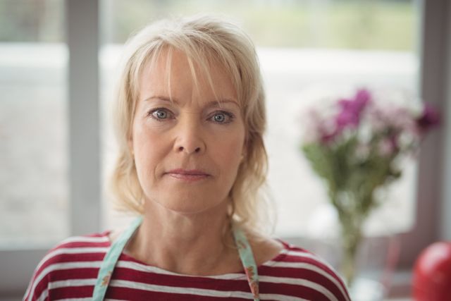 This image features a confident senior woman looking directly at the camera indoors. She is wearing a striped shirt and an apron, with natural light illuminating her face. The background includes blurred flowers, adding a homely touch. This image can be used for articles or advertisements related to senior lifestyle, health, confidence, and home living.