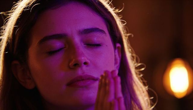 Young woman meditating with hands near chin in magenta and amber studio lighting closeup