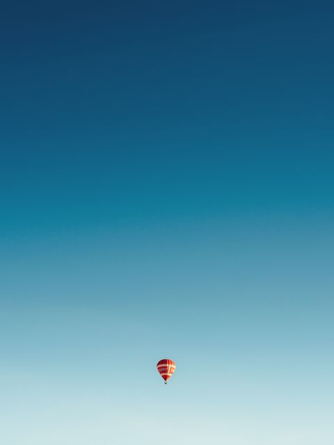 Hot air balloon floating in a clear, vast blue sky symbolizing freedom, adventure, and serenity. The minimalist composition accentuates the calm and solitude. Ideal for use in travel advertisements, inspirational posts, brochures, adventure themed marketing, and wallpapers to evoke a sense of wanderlust and tranquility.