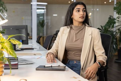 Indian businesswoman sitting at modern open-plan office desk holding notebook and laptop