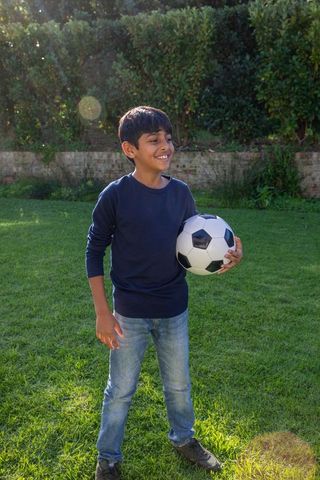 Smiling South Asian Boy Holding Soccer Ball on Green Lawn in Backyard