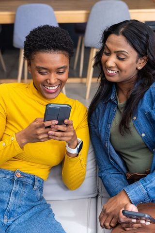 Smiling diverse women sharing smartphone on couch in cozy lounge, casual friendship