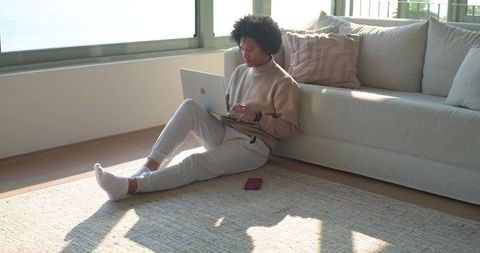 Sunlit african american woman working remotely on laptop by modern sofa