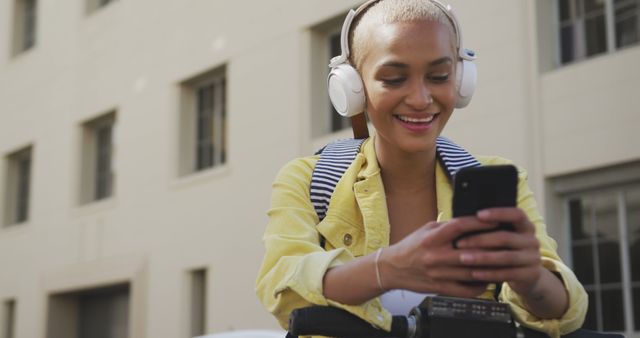 Woman wearing white headphones, checking her smartphone while standing outdoors. She is smiling and wearing a striped backpack. Ideal for campaigns related to modern communication, urban lifestyle, outdoor activities, and music streaming services.