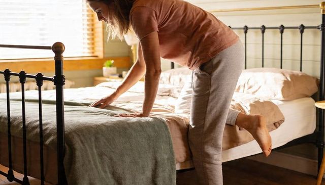Woman Smoothing Bed Sheets in Sunlit Cozy Bedroom with Metal Bed Frame and Green Throw