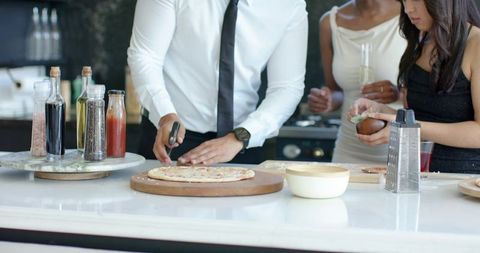 Friends in formal attire cutting pizza and toasting with champagne at kitchen counter