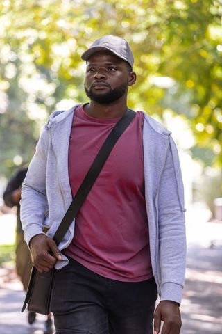 African man walking through sunlit urban park carrying crossbody bag and wearing casual cap