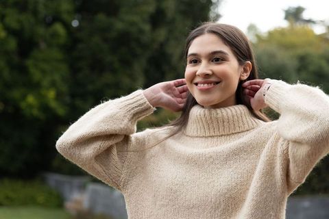 Smiling young woman adjusting cozy soft beige turtleneck sweater outdoors in leafy garden