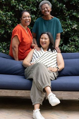 African american women laughing on navy couch in garden, showcasing smartwatch, friendship